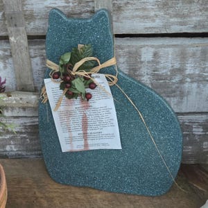 May include: A teal cat-shaped cutting board with a textured surface. It has a decorative bundle of faux berries and greenery tied with raffia. A white tag with text is attached to the board.