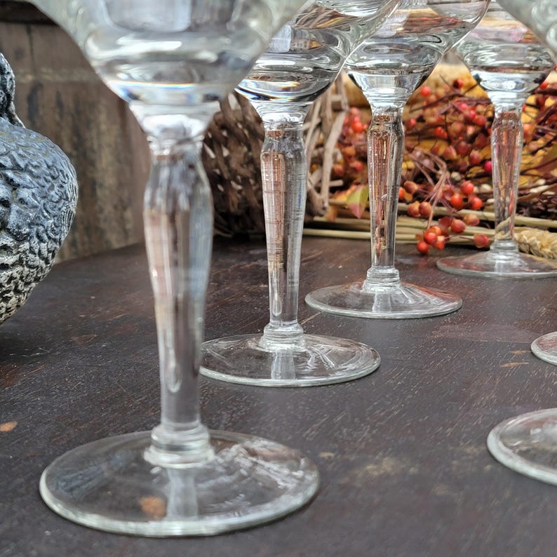 May include: A set of six clear glass martini glasses with a stem and a wide bowl. The glasses are arranged in a row on a dark wooden surface.