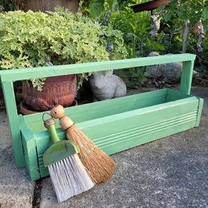 May include: A weathered, light green wooden garden tool caddy with a handle. A small green and white brush and a broom with a wooden handle rest against the caddy. A terracotta pot and a stone rabbit statue are in the background.