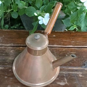 May include: A copper-colored metal vessel with a conical base, a flat lid featuring a small central knob, a short angled spout, and a long, slender wooden handle extending upward. The object has a smooth, metallic finish and a wide circular bottom.