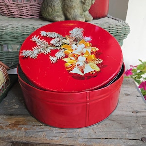 May include: A round red metal storage tin with a removable lid. The lid features a graphic illustration of white pine branches, gold ribbons, and three white bells with gold accents. The container has a smooth, solid red finish on the sides.