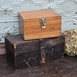 May include: A rectangular light brown wooden box with a silver metal latch sits atop a larger, weathered dark brown wooden box featuring a vertical metal strap closure and visible surface cracks on the front panel.