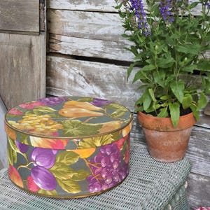 May include: A round hat box with a fruit pattern featuring grapes, pears, and plums in shades of purple, yellow, and red on a green background. The box has a gold trim and sits on a wicker surface. A potted plant with purple flowers is next to the box.