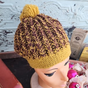 May include: A knitted beanie hat with a yellow pom-pom. The hat features a ribbed pattern in shades of mustard yellow and brown. The brim is a solid mustard yellow color.