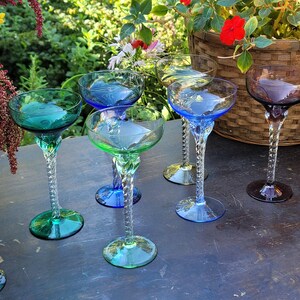 May include: A collection of six colorful, stemmed cocktail glasses. The glasses feature a twisted stem design and come in shades of green, blue, and purple. The glasses are arranged on a dark surface with a natural background.