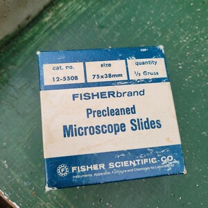 May include: A blue and white box of Fisher Brand precleaned microscope slides. The box has the text "FISHERbrand Precleaned Microscope Slides" and "FISHER SCIENTIFIC CO." printed on it. The box measures 75x38mm.
