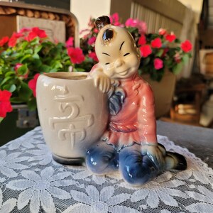 May include: A vintage ceramic planter in the shape of a smiling Asian boy, dressed in a red and blue outfit, leaning against a cream-colored pot. The pot has embossed characters. The planter is on a white lace tablecloth, with red flowers in the background.