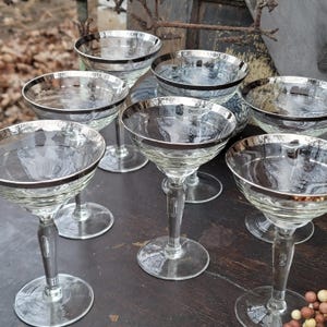May include: Set of eight vintage champagne glasses with silver rims. The glasses are clear and have a delicate design. They are arranged on a wooden surface.