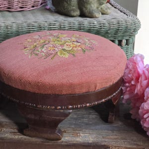 May include: A round footstool featuring a dusty pink needlepoint top with a central floral motif in shades of yellow, green, and cream. The base is constructed from dark brown wood with carved detailing along the edge and four short, sturdy legs.