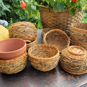 May include: A collection of small, woven baskets and terracotta pots. The baskets vary in shape and size, with a natural, light brown color. A large woven basket filled with greenery and red flowers is in the background. The items are arranged on a dark wooden surface.