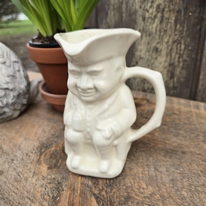 May include: Cream-colored ceramic pitcher in the shape of a seated man wearing a tricorn hat and coat. The handle is a curved branch shape. The pitcher is on a wooden surface.