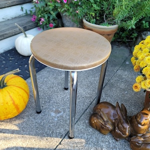May include: A vintage, round stool with a tan patterned seat and chrome legs. The seat is trimmed with white. The stool is a classic piece of furniture, suitable for various settings.