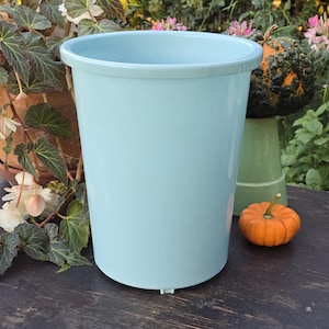 May include: A light blue cylindrical plastic flower bucket with a smooth finish and a reinforced rim. The container features a simple, open-top design with a flat base and small drainage-like notches at the bottom edge, presenting a solid color.