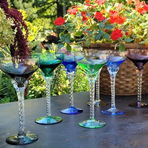 A set of 7 empty coupe or cocktail glasses is arranged on a dark wooden table outdoors. Each glass has a wide, shallow bowl and a long, twisted clear stem. The bowls come in different translucent colors—2 shades each of green & blue, 1 each of amber  smoky purple, gray. Foliage and red blossoms are visible in the blurred background, giving a garden or patio setting with soft natural light.