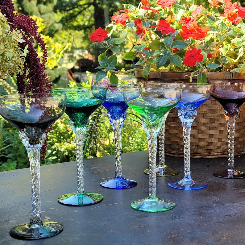 A set of 7 empty coupe or cocktail glasses is arranged on a dark wooden table outdoors. Each glass has a wide, shallow bowl and a long, twisted clear stem. The bowls come in different translucent colors—2 shades each of green & blue, 1 each of amber  smoky purple, gray. Foliage and red blossoms are visible in the blurred background, giving a garden or patio setting with soft natural light.