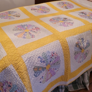 May include: A handmade quilt featuring a repeating fan pattern in a variety of pastel colors. The fans are set against a white background and framed by a bright yellow border. The quilt is placed on a bed.
