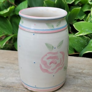 May include: A cylindrical ceramic vase with a cream-colored base, featuring hand-painted pink rose motifs and green leaves. The design includes thin horizontal pink and blue bands at the top and bottom, accented by small blue polka dots throughout.