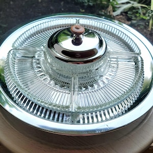 May include: A vintage serving dish with a clear glass insert and a shiny silver metal rim. The insert is divided into sections and has a ribbed design. A small, round, chrome-plated lid with a wooden knob sits in the center.