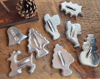 Puede incluir: Colección de cortadores de galletas navideñas de metal vintage en varias formas, incluyendo una campana, un árbol, un reno, un muñeco de nieve y una hoja de acebo. Los cortadores están dispuestos sobre una superficie de madera con una piña.