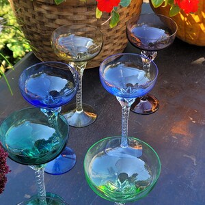 May include: A collection of six colorful champagne coupe glasses. Each glass features a unique bowl color, including blue, green, and yellow, with a twisted stem. The glasses are arranged on a dark surface, with a basket and flowers in the background.