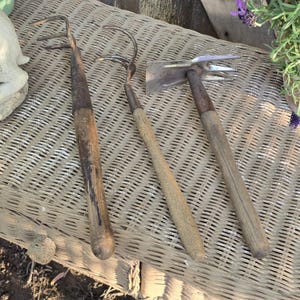May include: Three vintage gardening tools with wooden handles and metal heads rest on a woven surface. The tools include a hoe, a cultivator, and a weeding fork, showing signs of wear and use.