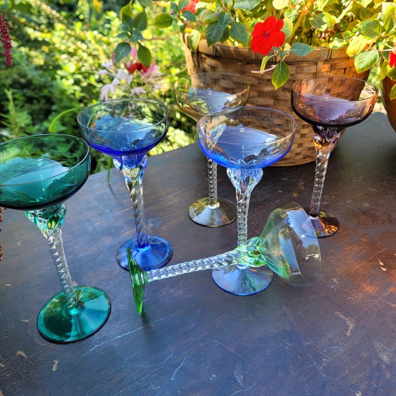 May include: A collection of colorful, vintage champagne coupe glasses. The glasses feature a twisted stem and a round bowl in shades of blue, green, and purple. The glasses are arranged on a dark surface, with a basket of flowers in the background.
