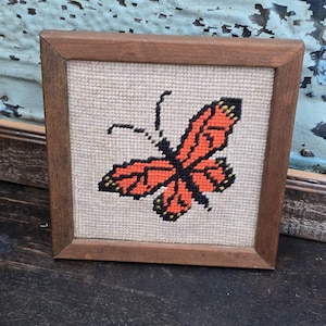 May include: A framed needlepoint featuring a butterfly. The butterfly has orange wings with black and yellow details, set against a beige background. The frame is made of brown wood, creating a vintage aesthetic.