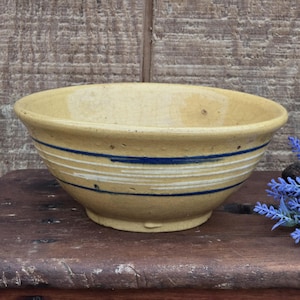 May include: A yellow ceramic bowl with blue and white horizontal stripes. The bowl has a rounded shape and a slightly flared rim. The bowl is sitting on a wooden surface.