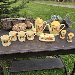 May include: A vintage cottage-themed tea set with a cream-colored base. The set includes a teapot, creamer, sugar bowl, butter dish, and six mugs. Each piece features a charming cottage design with red roofs and green trees, evoking a rustic aesthetic.