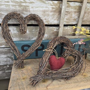 May include: Two rustic heart-shaped wreaths made of intertwined brown twigs. One large heart stands upright, while the other has a small, red felt heart inside. The wreaths are displayed on a wooden surface with a weathered background.