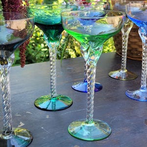 May include: A collection of colorful, vintage-style champagne glasses. Each glass features a unique swirl stem and a colored bowl in shades of green, blue, and yellow. The glasses are arranged on a dark surface, showcasing their vibrant hues.