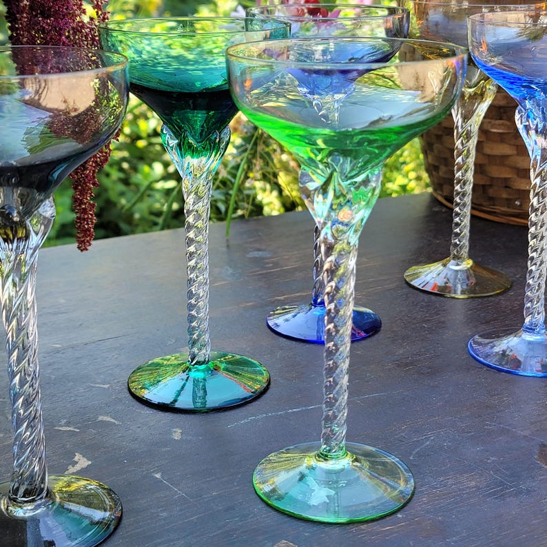 May include: A collection of colorful, vintage-style champagne glasses. Each glass features a unique swirl stem and a colored bowl in shades of green, blue, and yellow. The glasses are arranged on a dark surface, showcasing their vibrant hues.