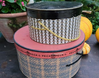 May include: Two vintage hat boxes stacked on top of each other. The top box has a black lid and a geometric pattern in white, brown, and gold. The bottom box is pink with a blue and tan patterned design and the text "PECK & PECK".