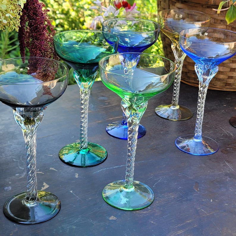 May include: A collection of colorful, stemmed champagne glasses. Each glass features a unique hue, including blue, green, and yellow, with a twisted stem design. The glasses are arranged on a dark surface, suggesting a celebratory or decorative theme.