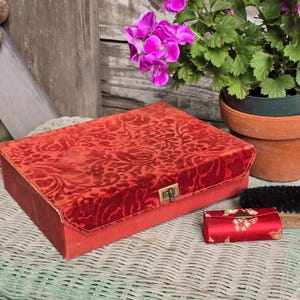 May include: A rectangular red velvet box with a floral pattern and a gold-tone metal latch, accompanied by a smaller, matching red fabric case with a gold floral print and a gold-tone metal clasp, both resting on a textured surface.