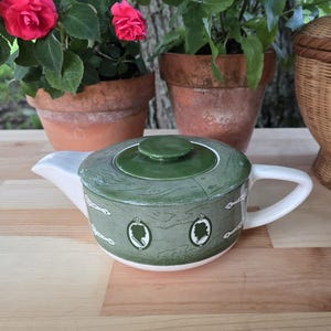 May include: A ceramic teapot featuring a sage green body with a wood grain pattern, a solid dark green lid, and a white handle and spout. The exterior is decorated with white oval cameos displaying silhouettes and small white decorative accents.