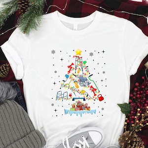 May include: White t-shirt with a Christmas tree design made up of medical equipment, including crutches, a walker, a wheelchair, and a weight. The tree is decorated with lights and ornaments. The t-shirt is on a red and black plaid blanket with a gray beanie, a pair of gray sneakers, and a small pine cone.