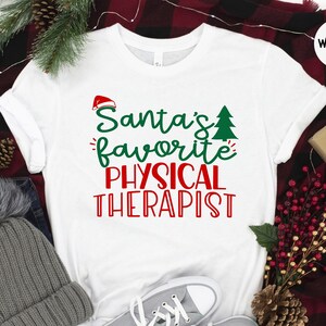 May include: White t-shirt with red and green text that reads "Santa's favourite physical therapist". The shirt is on a red and black checked blanket with a grey beanie, pine cones, and red berries.