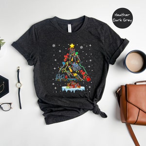 May include: A dark heather grey t-shirt with a Christmas tree design made of tools. The tree is decorated with ornaments and a star on top. The text "Heather Dark Grey" is visible in the upper right corner of the image.