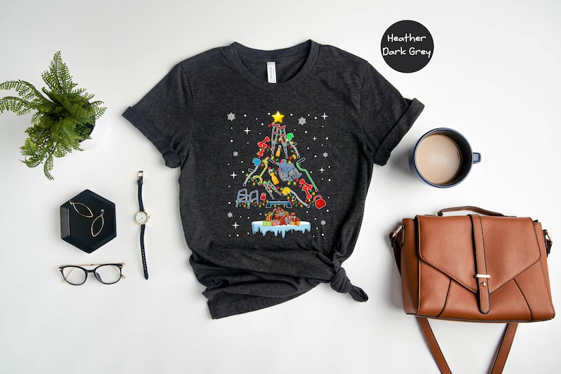 May include: A dark heather grey t-shirt with a Christmas tree design made of tools. The tree is decorated with ornaments and a star on top. The text "Heather Dark Grey" is visible in the upper right corner of the image.