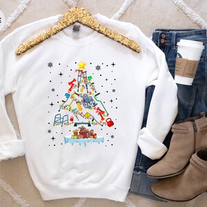 May include: White sweatshirt with a Christmas tree design made of various medical equipment, including crutches, a wheelchair, and a walker. The tree is decorated with ornaments and a star on top. The sweatshirt is on a white background with a pair of brown boots and blue jeans.