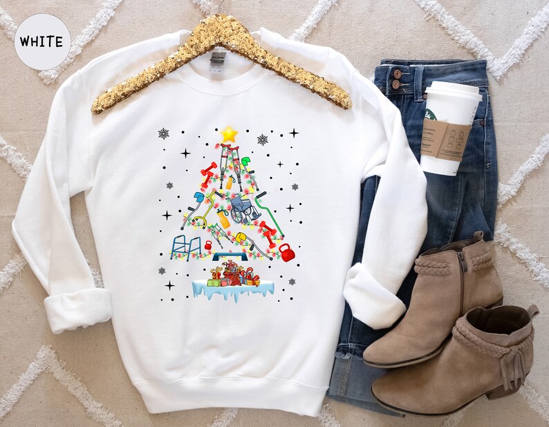 May include: White sweatshirt with a Christmas tree design made of various medical equipment, including crutches, a wheelchair, and a walker. The tree is decorated with ornaments and a star on top. The sweatshirt is on a white background with a pair of brown boots and blue jeans.