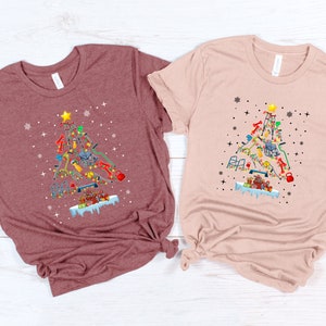 May include: Two t-shirts with a Christmas tree design made of fitness equipment. The tree is decorated with ornaments and a star on top. The t-shirts are in maroon and light pink.