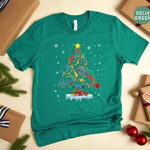 May include: A teal green t-shirt with a Christmas tree design made of fitness equipment. The tree is decorated with ornaments and lights. The text "Kelly Green" is visible on a brown gift box in the background.