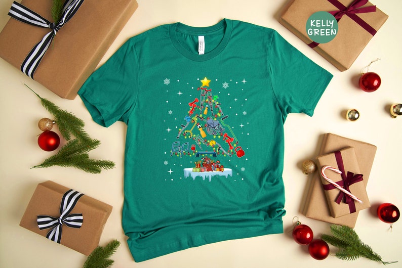 May include: A teal green t-shirt with a Christmas tree design made of fitness equipment. The tree is decorated with ornaments and lights. The text "Kelly Green" is visible on a brown gift box in the background.