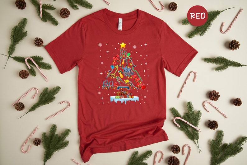 May include: Red t-shirt with a Christmas tree design made of fitness equipment. The tree is decorated with lights and ornaments. The text "RED" is in the upper right corner of the image.