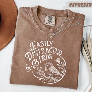 a brown t-shirt with a white graphic design, a brown hat, and some dried leaves, all placed on a white surface.