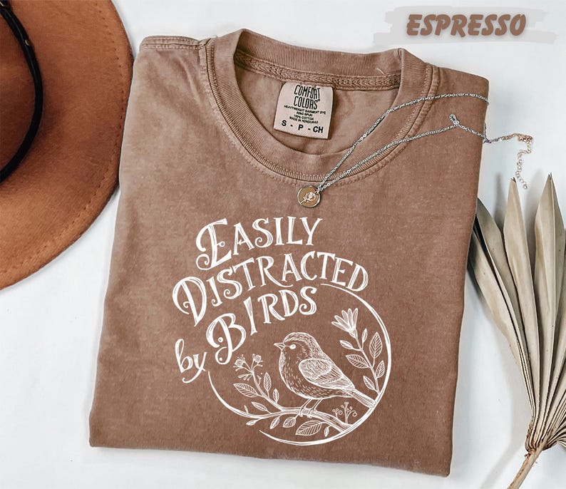 a brown t-shirt with a white graphic design, a brown hat, and some dried leaves, all placed on a white surface.