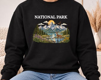 Może przedstawiać: Czarna bluza z okrągłym dekoltem z nadrukiem graficznym z napisem NATIONAL PARK. Ilustracja przedstawia pasmo górskie, jezioro, jelenia stojącego w wodzie, sosny, fioletowe kwiaty oraz żółte słońce na niebie.