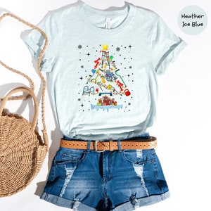 May include: A light blue t-shirt with a Christmas tree design made up of various fitness equipment. The tree is decorated with lights and presents. The shirt is displayed on a model with denim shorts and a straw bag.